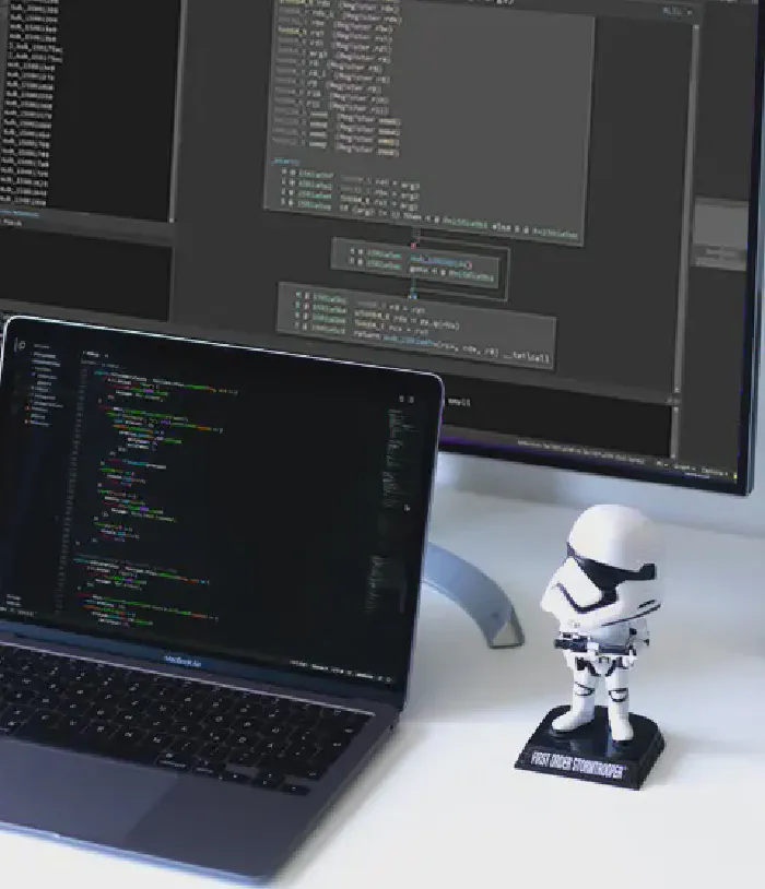 Laptop with code, monitor with binary ninja, small storm trooper toy in the front