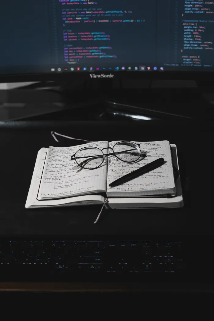 Keyboard, notebook with glasses on it, large computer screen in the back